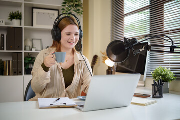 Young woman podcaster smiling and holding a coffee mug while recording a podcast in a modern home...