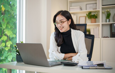 Fototapeta premium Confident businesswoman in glasses smiling during a video call while working on laptop in a modern office.