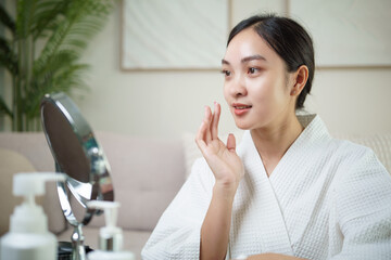 Fototapeta premium Young Asian woman applying skincare cream in front of a mirror. Wellness and self care concept.