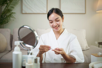 Smiling woman in bathrobe applying skincare in front of mirror during her morning routine. Wellness...