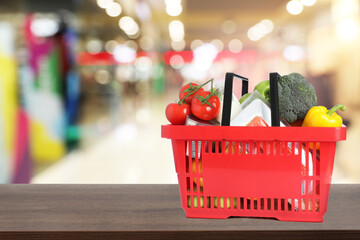 Red shopping basket full of groceries on wooden table in supermarket. Space for text