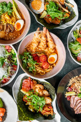 A vibrant assortment of Asian cuisine featuring ramen with tempura shrimp, fresh salads, seafood, and vegetables. The visually appealing dishes showcase a variety of textures, colors