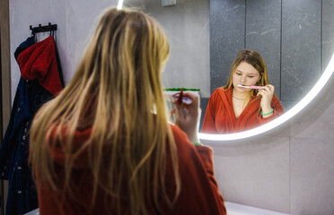 Young woman in a red robe stands at the sink brushing her teeth, focused on her reflection in a...