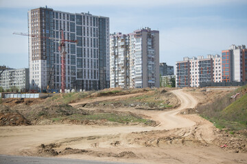 construction of the foundation of a new residential building using a tower crane in a large neighborhood with new tall modern multi-storey buildings on the background of which a winding dirt road lead