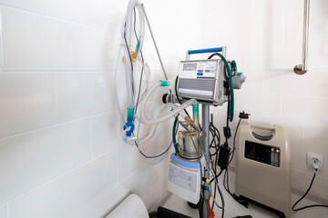 A ventilator, a complex medical apparatus with tubing and controls, stands next alongside a portable oxygen concentrator and to a hospital bed in a clean, tiled room.