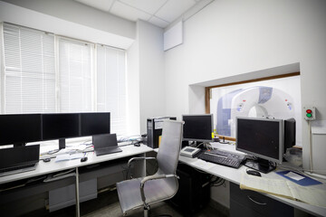 A medical diagnostic control room features multiple computer monitors, laptops, and an office chair, with a window view into an adjacent CT scanner room on the basement floor of the facility.