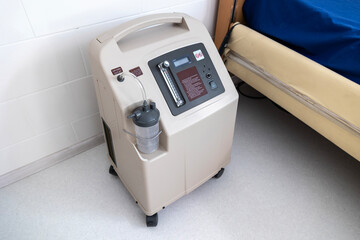 A portable oxygen concentrator stands on a tiled floor next to a hospital bed, showcasing its controls and humidifier bottle in a medical room.