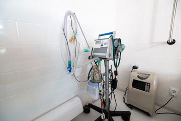 A ventilator, a complex medical apparatus with tubing and controls, stands next alongside a portable oxygen concentrator and to a hospital bed in a clean, tiled room.