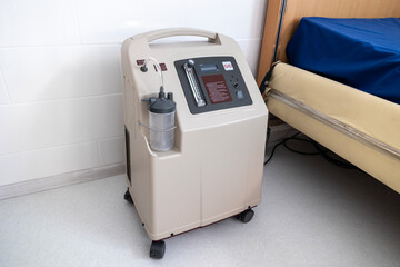 A portable oxygen concentrator stands on a tiled floor next to a hospital bed, showcasing its controls and humidifier bottle in a medical room.