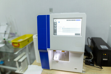 A medical blood analyzer with a digital display showing analysis results and a black uninterruptible power supply sit on a wooden table in a clinic room in a clinic hospital.