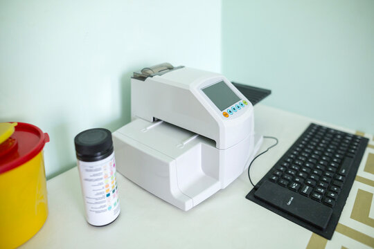 A white urine analyzer with a display and a black keyboard sits on a white table next to a container of urine test strips.