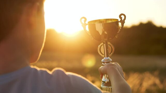 Child hand holding trophy at sunset symbolizes success, motivation, and achievement. Powerful image of determination, motivation, childhood dreams, achievement and personal victory under golden light.