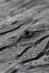 A silver ring with a square black stone rests delicately on a dramatic, cracked surface that resembles volcanic rock. The scene evokes mystery, strength, and timeless elegance.

