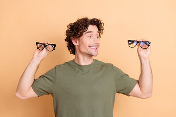 Joyful young man in casual outfit holding stylish glasses in each hand against a beige background