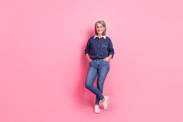 Confident mature woman standing casually against a pink background, showcasing her stylish outfit...