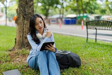 Young female student is sitting under a tree in a park, taking notes on her tablet and enjoying the...