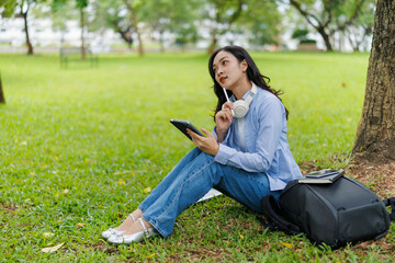 Female college student sitting under a tree in a park, using a tablet, holding a pen and wearing headphones around her neck, thinking about a project