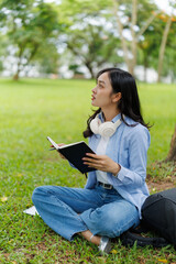 Fototapeta premium Asian female university student holding a notebook and pen is sitting under a tree on the grass in a park, taking notes and learning outdoors