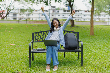 Young Asian student celebrating success with arms raised while using a laptop, sitting on a bench in a vibrant green park, embodying joy and achievement in her educational journey