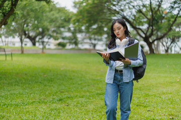 Fototapeta premium Young woman with headphones around her neck and backpack is reading a book while walking in a green park, she is carrying a laptop with her and enjoying the fresh air