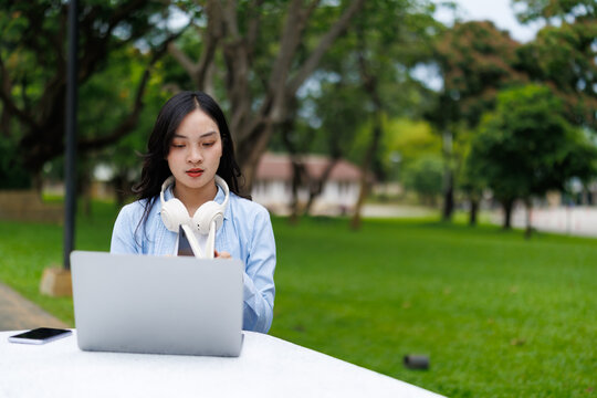 Concentrating young Asian freelancer working on a laptop while using a smartphone in a park, enjoying the fresh air and the beauty of nature in a serene outdoor setting