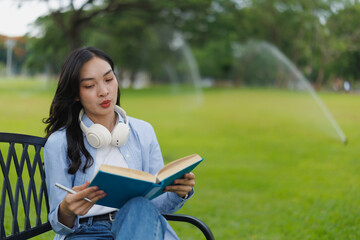 Obraz premium Asian student reading a book while sitting on a park bench, holding a pen and enjoying the peaceful atmosphere as automatic sprinklers water the lush green grass nearby