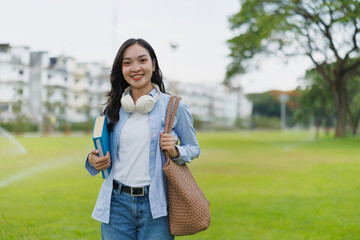 Young happy asian female student is walking in a park holding books and a bag while wearing headphones around her neck, returning from university after classes