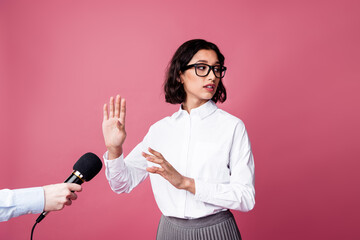Charming young woman expressing disinterest in an interview while standing against a vibrant pink...