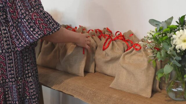 Woman carefully tying red ribbons on burlap gift bags, decorative flower arrangement beside handcrafted packaging on weathered wooden surface