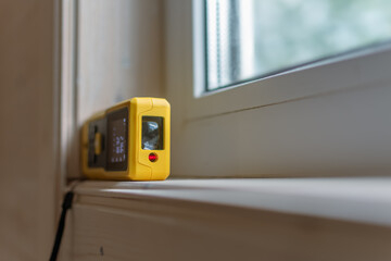 Yellow laser distance measurer placed on a window sill, its digital screen illuminated, ready to...