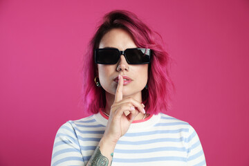 Fototapeta premium Beautiful woman with bright dyed hair in sunglasses making hush gesture on pink background