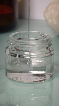 Transparent gel-like liquid being poured into a glass jar against a blue glossy background. Skincare or cosmetic product preparation. Clean, modern, and minimalistic beauty concept.