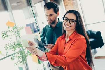 Smiling colleagues in stylish office collaborating on startup project, using sticky notes on transparent board.