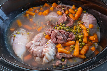 Close-up image of raw ingredients in a slow cooker, featuring chunks of beef, whole chicken thighs, baby carrots, and peas