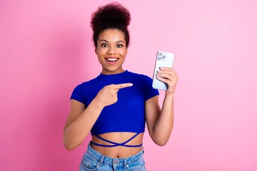 Gordijnen Excited young woman holding a smartphone in hand while pointing at it against a pink background © deagreez