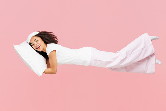 Fototapeta Full body side view calm young woman wearing pyjamas jam sleep eye mask rest relax at home fly fall hover over air hurry up hold pillow isolated on plain pink background. Good mood night nap concept.