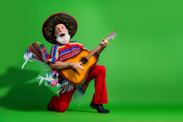Elderly man in colorful Mexican attire strumming a guitar against a vibrant green backdrop...