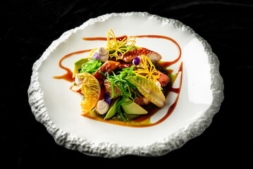 A gourmet dish elegantly plated with fresh vegetables, citrus slices, meat, and edible flowers, presented on a textured white plate against a dark background, highlighting vibrant colors