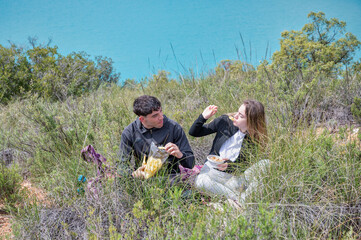 Teen girl and boy having a picnic after hiking, sitting in the grass and enjoying food together while relaxing beside a turquoise lagoon in a peaceful natural setting