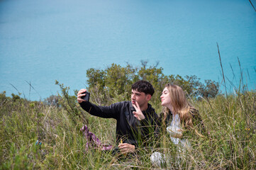 Teen girl and boy sitting in the grass after a hike, smiling and taking a selfie together with a turquoise lagoon in the background on a calm and joyful outdoor break