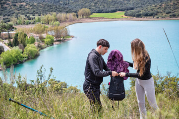 Teen girl and boy organizing their backpack together during a hiking break, folding a jacket with focus and teamwork beside a turquoise lagoon and natural landscape