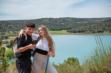 Teen girl and boy hiking together on a hillside trail by a turquoise lagoon, checking a smartphone and smiling on a sunny outdoor day