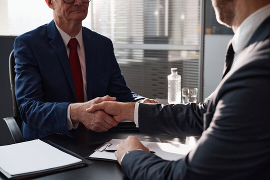 Over shoulder shot of senior Caucasian man in suit shaking hands with male colleague making business deal, while sitting at desk during business meeting in modern office - Powered by Adobe