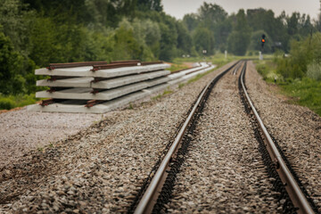 Fototapeta premium Long, straight railway tracks stretch into the distance between green forest areas under a cloudy sky, offering strong perspective and symmetry.