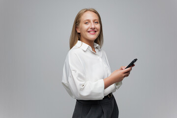 Cheerful Young Woman Smiles While Texting on Her Smartphone Against a Simple Gray Backdrop,...
