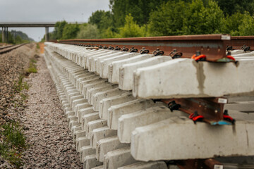 A close side view of stacked concrete railway track sections alongside a railway line, with a bridge visible in the background under cloudy skies.