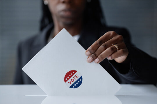 Cropped shot of Black female election participant holding envelope with vote sticker, placing completed ballot into voting box at polling station