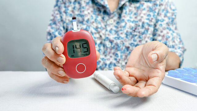 Senior woman holding red glucometer showing 145 mg/dL blood sugar level after fingertip test demonstrating diabetes monitoring at home - Powered by Adobe