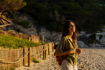 Young woman with backpack walking along a fenced path on a beautiful beach at sunset, enjoying summer vacation