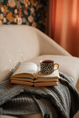 Cup of coffee staying on stack of open paper books and knitted sweater on couch in living room at...